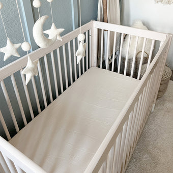 A beige crib with a light beige fitted sheet is centered in the image. The crib has wooden slats and a simple, clean design. A felt moon and star mobile hangs over the crib. The mobile's components are also beige and white. A light beige/cream colored wall is behind the crib. A woven basket and other light-colored decorative items are partially visible in the background. A light-colored rug is visible underneath the crib.