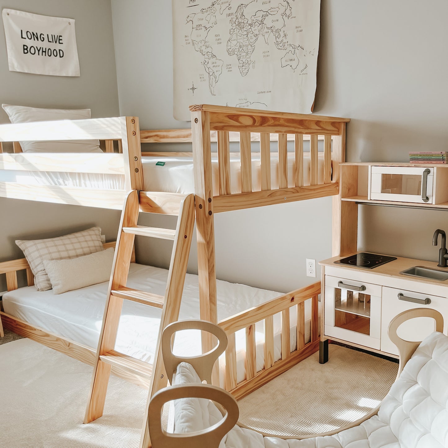 A bright kids' room features wooden bunk beds topped with a Newton Baby Waterproof Organic Twin Mattress Pad, a play kitchen, beige pillows, world map wall art, and a "LONG LIVE BOYHOOD" sign—all in a light, neutral color scheme.