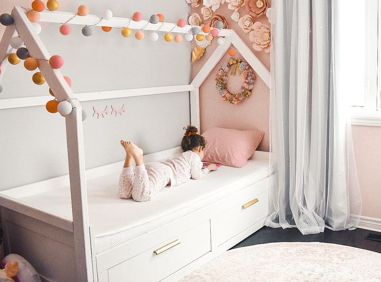 A young child relaxes on a house-shaped bed adorned with colorful pompoms and floral decors in a cozy, pink-themed bedroom.