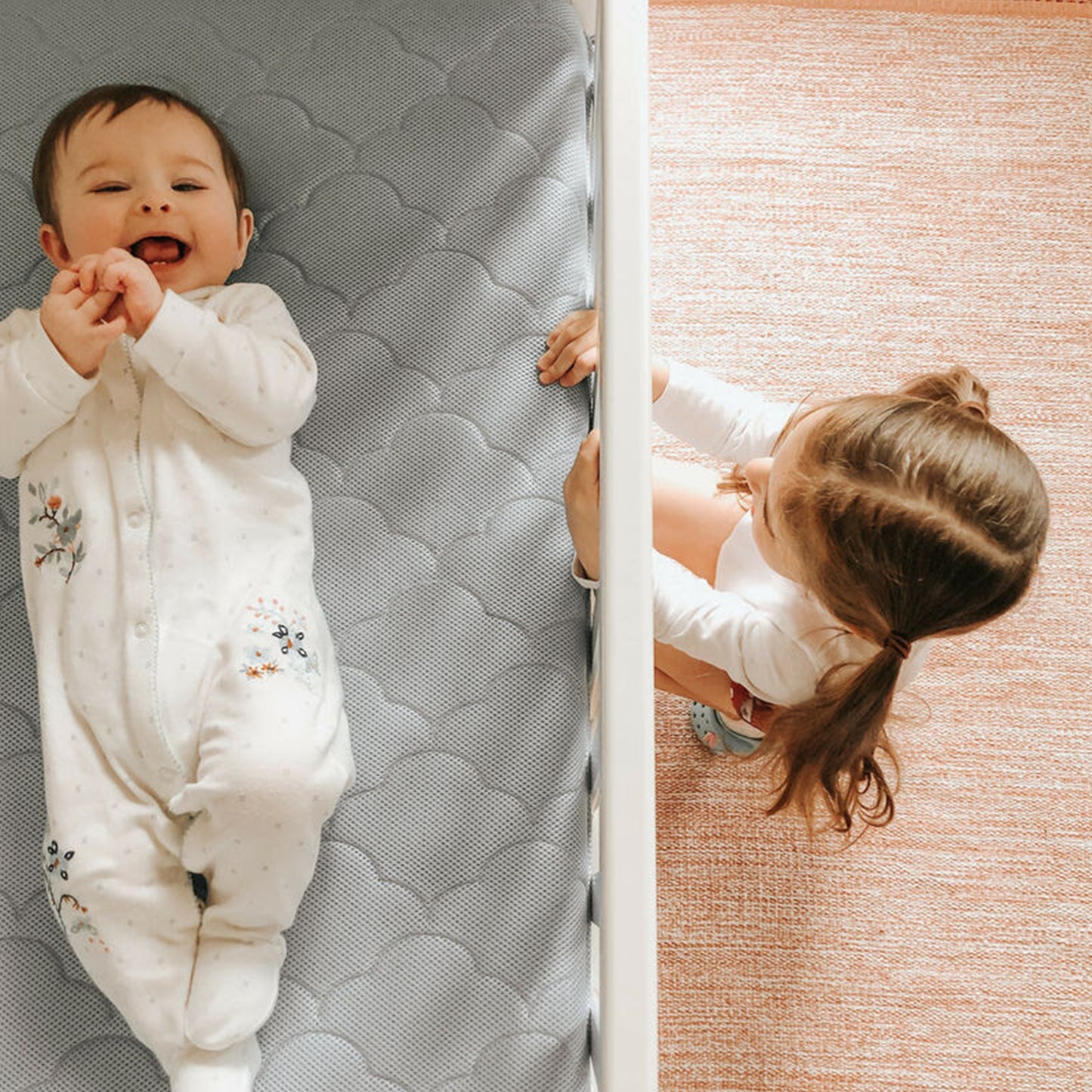 baby lying on back on mattress laughing with sister peering through crib at baby