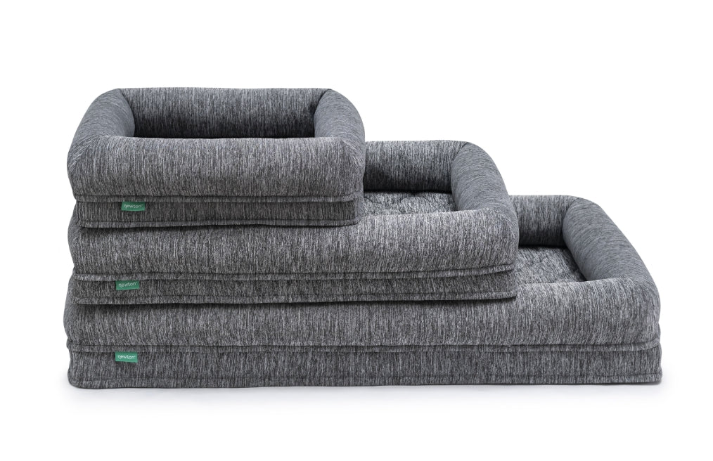 Three dog beds, stacked in descending order of size, are displayed against a white background. The beds are a dark gray or charcoal color with a slightly textured, woven appearance.  Each bed has a raised, slightly padded edge. Small, green rectangular labels are visible on the seams of each bed. The beds are oriented in a way that displays their sides and the stacked arrangement is apparent. The beds appear to be made of a soft, comfortable material.