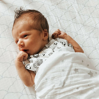 A newborn baby, with light brown hair, is lying on a white changing pad or blanket. The blanket has a light gray, scalloped pattern resembling clouds. The baby is wearing a short-sleeved bodysuit with a black and white animal print, including images of a zebra, rhinoceros, and an elephant. A white, star-printed receiving blanket covers the baby partially. The baby's eyes are open, and they are looking slightly to the right of the image. One hand is raised to the mouth. The baby appears calm and relaxed.