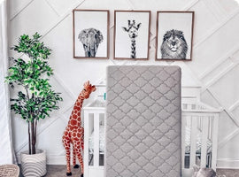 A nursery with white diamond-patterned wainscoting displays three black-and-white animal prints—elephant, giraffe, and lion. A plush faux-fur giraffe stands beside a white crib, while a gray quilted mattress rests in front. A leafy plant in a beige pot adds warmth, with a cozy basket and gray stuffed toy peeking from the floor.