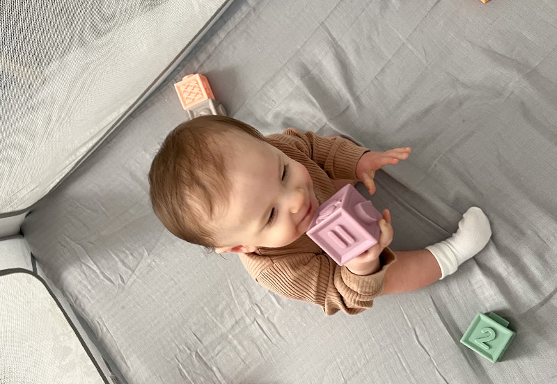A baby in a ribbed brown outfit and white socks sits on Travel Crib & Play Yard Sheets, holding and chewing a purple block while colorful blocks are scattered nearby.