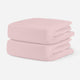 Light pink, breathable cotton crib sheets stacked neatly on a plain background. skip