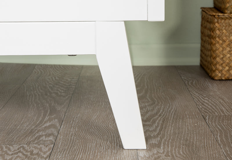 Close-up of a white wooden leg of the Soho Convertible Crib by dadada baby on a gray wood floor, with part of a woven basket in the background—highlighting clean lines and timeless mid-century style.