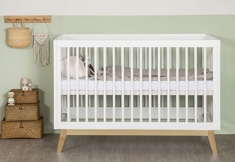 Modern white baby crib in a serene nursery setting with green walls, styled with wicker storage baskets and a decorative wall hanging.
