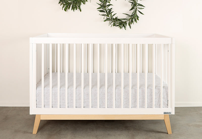 The dadada baby Soho Convertible Crib, a modern white 3-in-1 crib with light wood legs and a patterned mattress sheet, boasts mid-century style and sits against a white wall adorned with two green leafy wreaths.