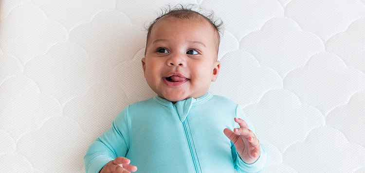 A smiling baby in a light blue zip-up onesie lies on a white, textured surface, looking up with their tongue slightly out and hands raised.