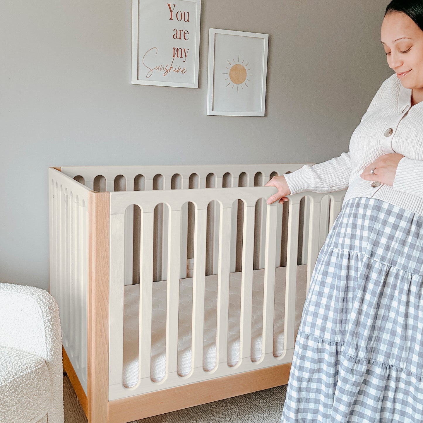 A pregnant woman stands beside a Newton Galileo Convertible Crib by Newton Baby in a softly lit nursery, gently touching her belly. Above the crib, wall art reads "You are my sunshine" with a sun illustration.