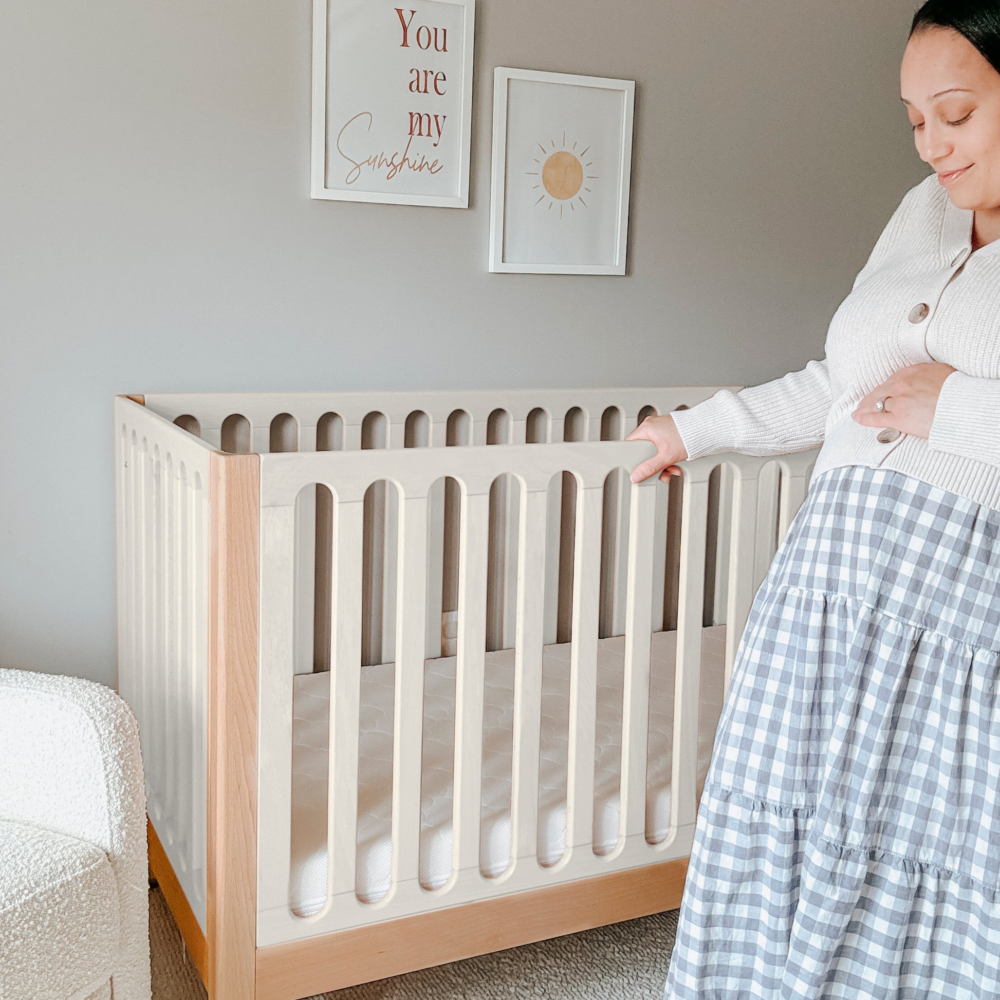A pregnant woman stands beside a Newton Galileo Convertible Crib by Newton Baby in a softly lit nursery, gently touching her belly. Above the crib, wall art reads "You are my sunshine" with a sun illustration.