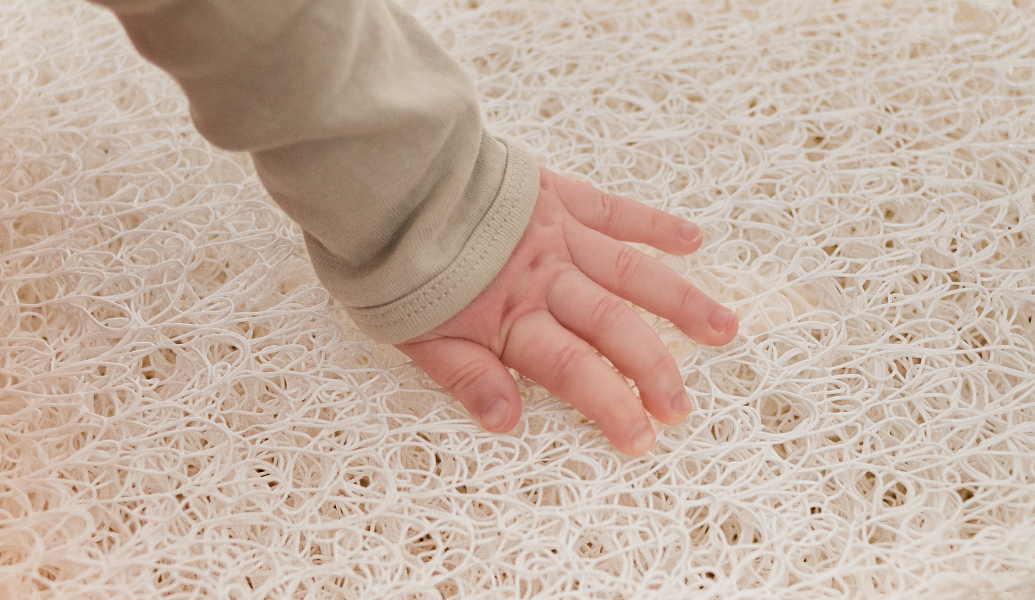 A delicate, lace-like web of cream-colored fibers stretches across the frame, its porous weave creating an intricate pattern. A smooth, beige hand rests lightly atop the surface, fingers gently splayed—just enough to reveal the material’s airy texture without disturbing its structure. The interplay of light and shadow highlights the delicate craftsmanship of the woven strands.