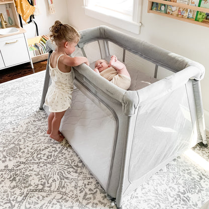 A young girl smiles beside a Deluxe Travel Crib & Play Yard by Newton Baby, where a baby lies inside on a breathable crib mattress. The bright room features white walls, a patterned rug, and ample natural light from the window.
