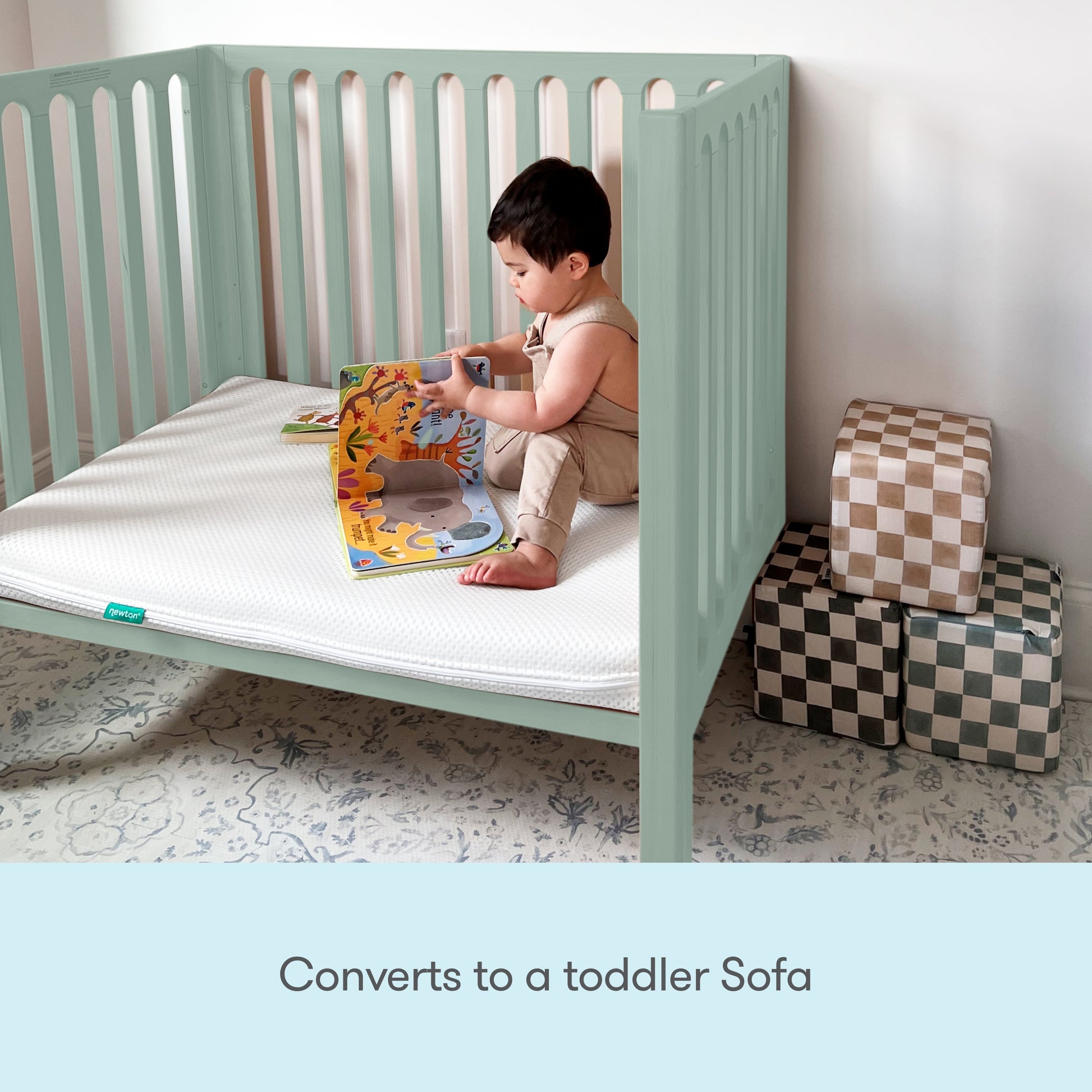 A young child sits in a mint green Newton Galileo Mini Crib with Newton Mattress, now converted to a toddler sofa, reading a colorful book. Three checkered storage cubes are stacked nearby. Text below reads: Converts to a toddler sofa.