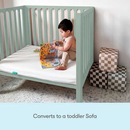 A young child sits in a mint green Newton Galileo Mini Crib with Newton Mattress, now converted to a toddler sofa, reading a colorful book. Three checkered storage cubes are stacked nearby. Text below reads: Converts to a toddler sofa.