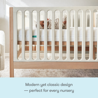 A baby lies in the Newton Galileo Convertible Crib by Newton Baby, featuring modern beech wood and white slats. Modern yet classic design—perfect for every nursery, and easily converts with our toddler bed conversion kit.
