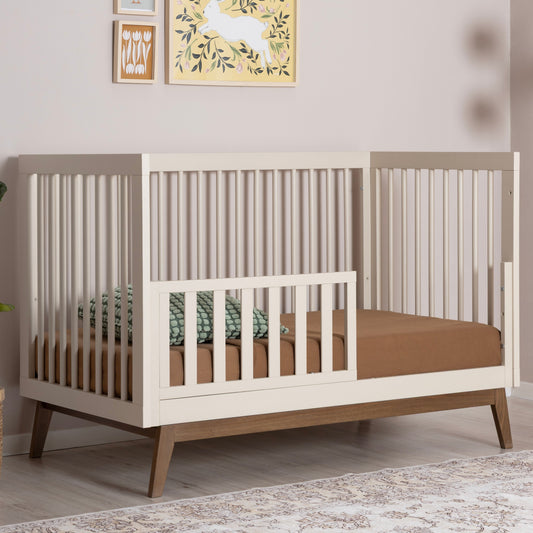 A modern dadada baby crib with white slats, brown legs, and a Crib Conversion Kit (Toddler Bed Rail) on one side, styled with a brown mattress, green pillow, and set against a beige wall decorated with artwork.