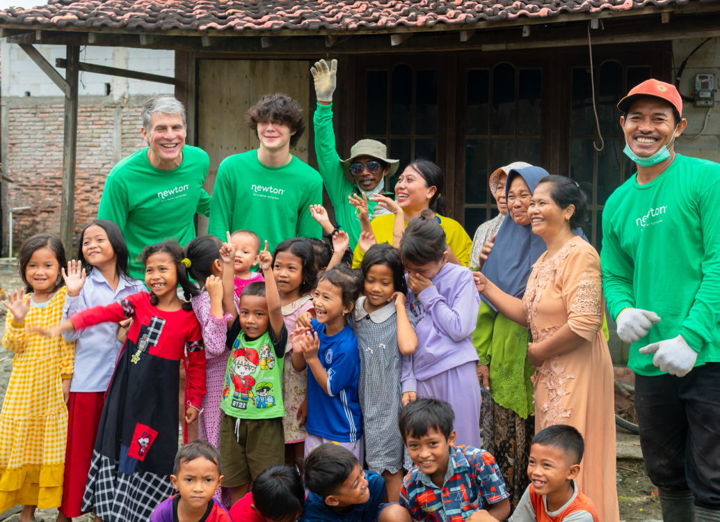 A lively group of adults and children from a rural area gather outside a simple wooden building. Volunteers in lime-green "Newton" shirts mingle with women in traditional headscarves, while children in colorful clothes cheer with raised hands. The scene radiates joy and togetherness, capturing the spirit of a shared celebration or volunteer initiative. The modest wooden structure and rural backdrop emphasize the close-knit, communal atmosphere.