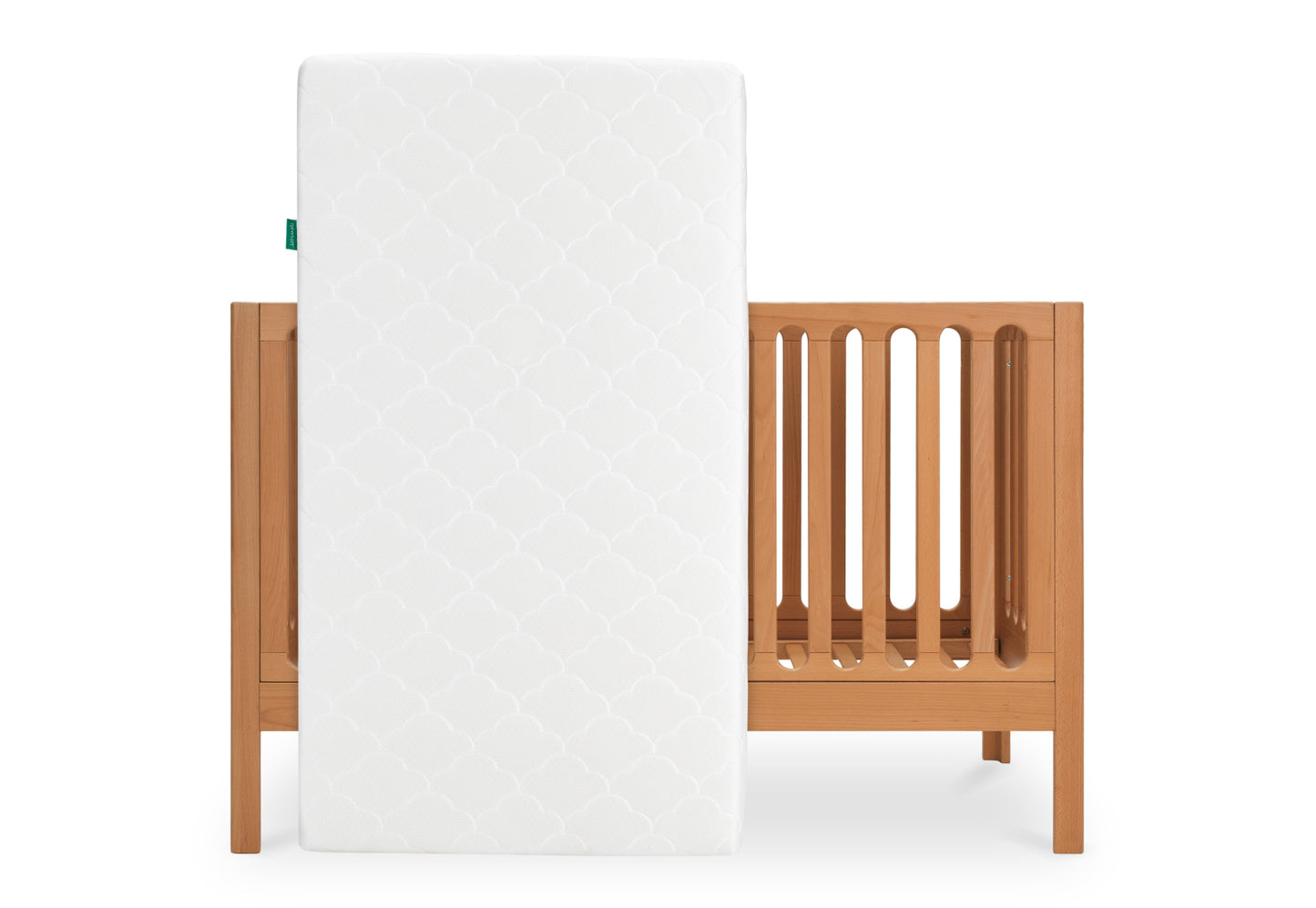 White crib mattress with a scalloped pattern standing next to a wooden crib on a white background.