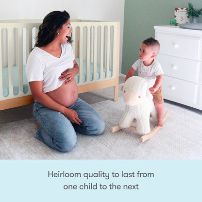 A pregnant woman smiles as a toddler rides a plush rocking sheep in a cozy nursery, featuring the Newton Galileo Convertible Crib by Newton Baby. Text reads: Heirloom quality to last from one child to the next.