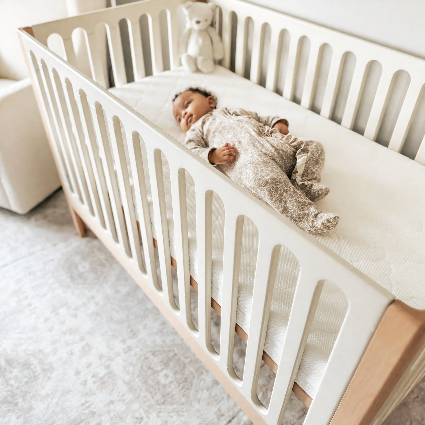 A baby in patterned pajamas lies on their back in a Newton Galileo Convertible Crib by Newton Baby, featuring white rails and a beech wood frame. A white stuffed bear sits in the corner, with a soft glow and light-colored rug completing the scene.