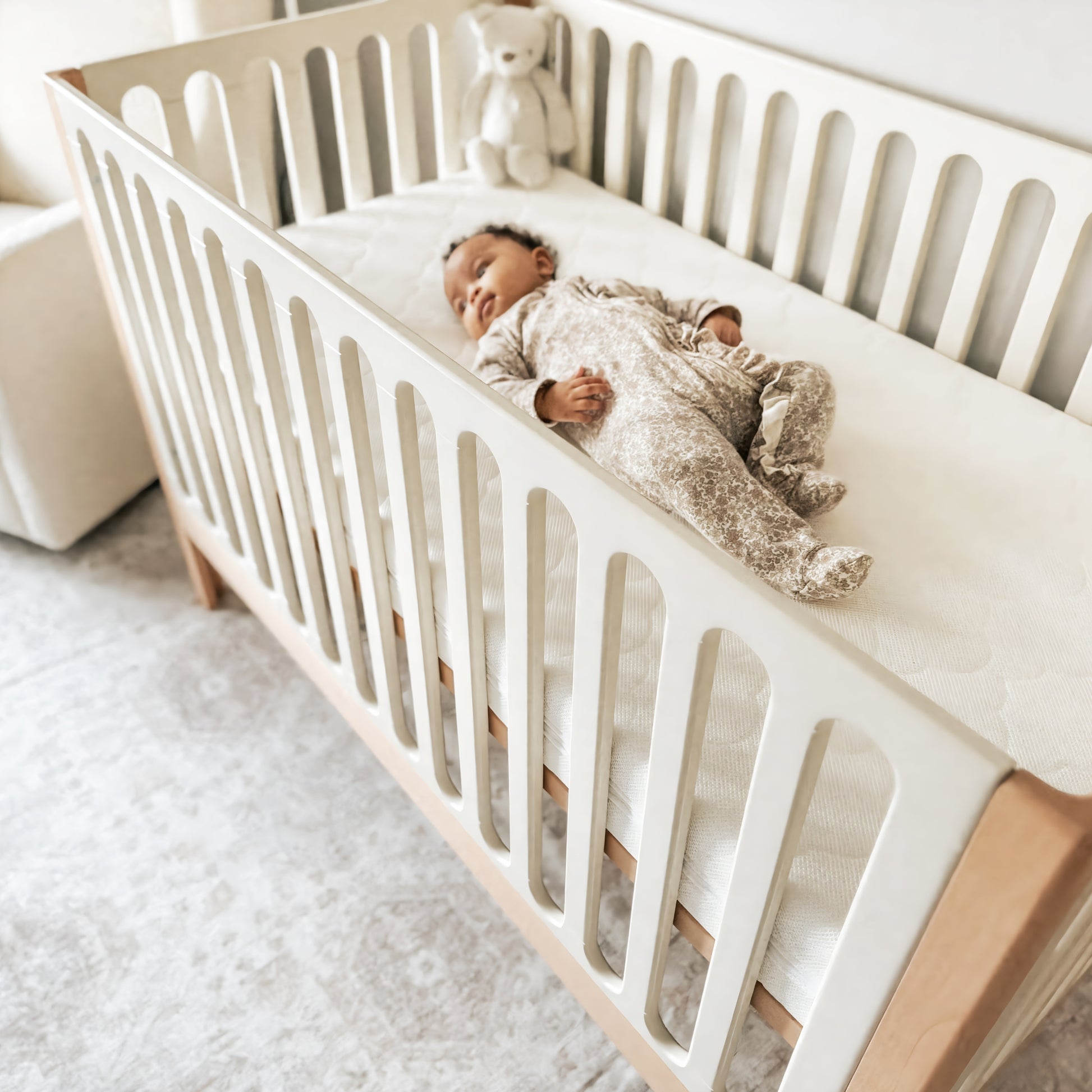 A baby in patterned pajamas lies on their back in a Newton Galileo Convertible Crib by Newton Baby, featuring white rails and a beech wood frame. A white stuffed bear sits in the corner, with a soft glow and light-colored rug completing the scene.