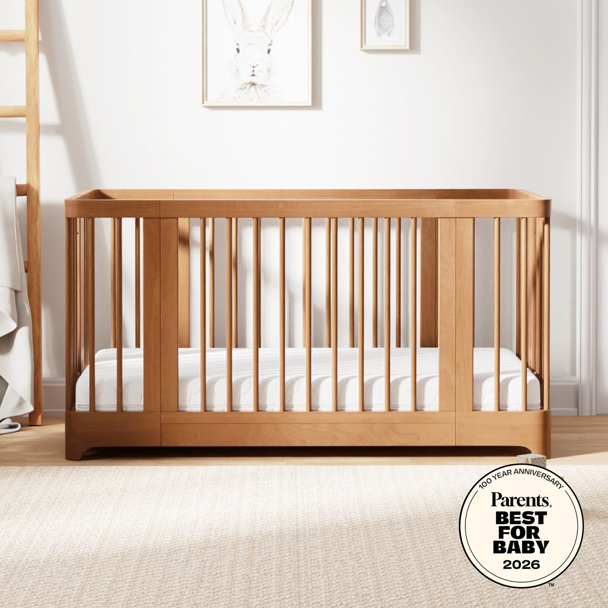 A Newton Baby Newton Nest Convertible Crib with vertical slats and a striped mattress sits in a softly lit nursery. Animal art hangs above, and the crib features GREENGUARD Gold certification and a Parents Best for Baby 2026 award badge. 999-8442-CST-C1