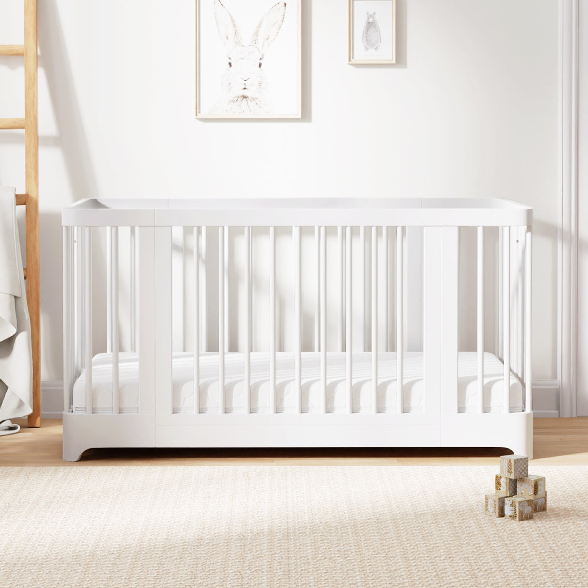 A modern Newton Nest Convertible Crib by Newton Baby, GREENGUARD Gold certified and featuring slatted sides in a white finish, sits on a beige carpet. Three wooden toy blocks are stacked nearby. Art featuring a rabbit and a smaller framed picture hang on the wall above the crib. 999-8442-WHT-C1