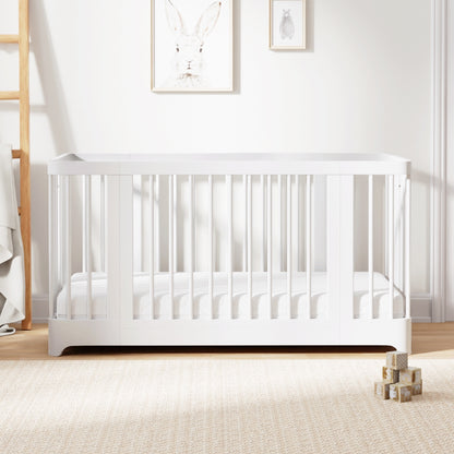 A modern Newton Nest Convertible Crib by Newton Baby, GREENGUARD Gold certified and featuring slatted sides in a white finish, sits on a beige carpet. Three wooden toy blocks are stacked nearby. Art featuring a rabbit and a smaller framed picture hang on the wall above the crib. 999-8442-WHT-C1