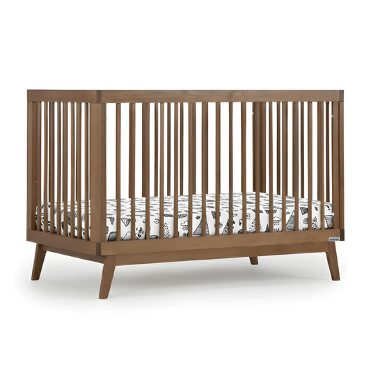 The dadada baby Soho Convertible Crib features vertical slats and angled legs paired with a white Newton crib mattress decorated with black line art, showcasing a sleek mid-century style in a versatile 3-in-1 design. 500-8220-WAL
