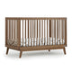 The dadada baby Soho Convertible Crib features vertical slats and angled legs paired with a white Newton crib mattress decorated with black line art, showcasing a sleek mid-century style in a versatile 3-in-1 design. 500-8220-WAL
