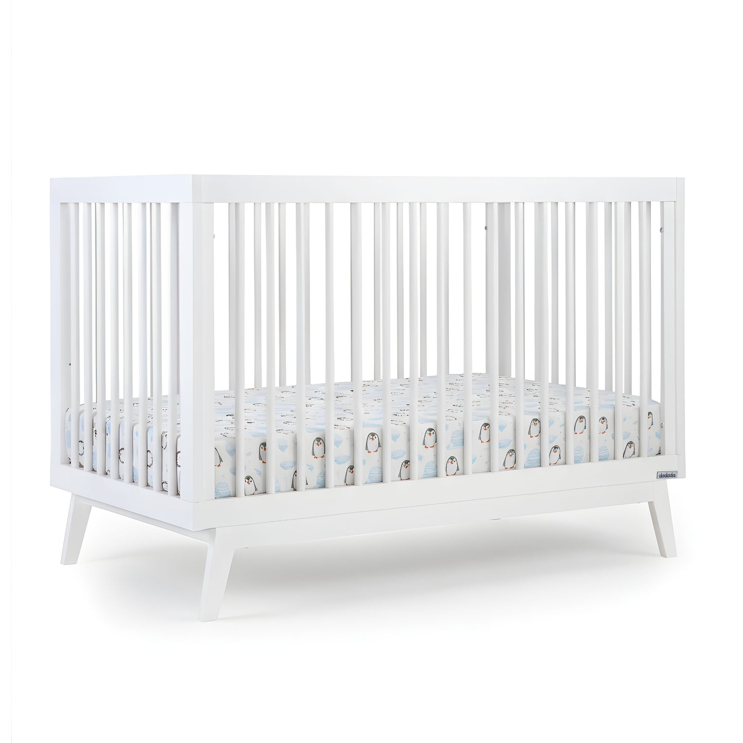 The dadada baby Soho Convertible Crib, a modern white 3-in-1 crib with vertical slats and mid-century angled legs, is shown with a Newton crib mattress and a fitted sheet featuring a cute penguin pattern on a plain white background.