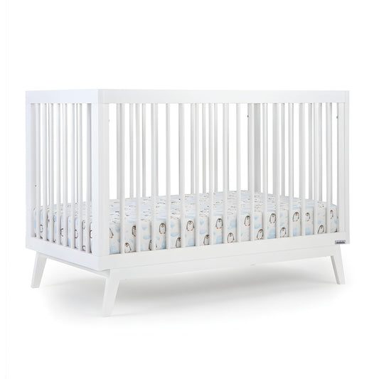 The dadada baby Soho Convertible Crib, a modern white 3-in-1 crib with vertical slats and mid-century angled legs, is shown with a Newton crib mattress and a fitted sheet featuring a cute penguin pattern on a plain white background.