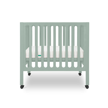 The Newton Galileo Mini Crib with Newton Mattress by Newton Baby is shown in light green with vertical slats, a white mattress, and four black wheels against a plain white background.