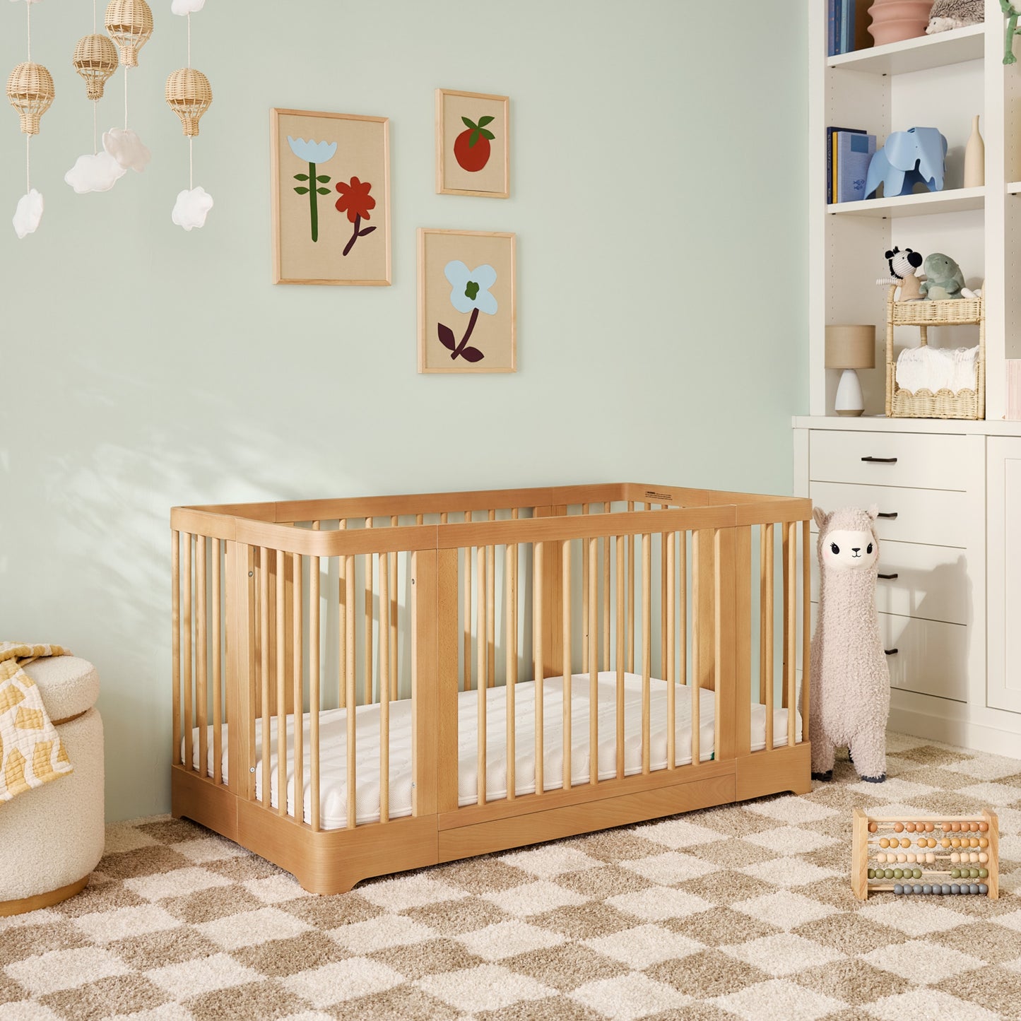 A nursery featuring the Newton Nest Convertible Crib by Newton Baby in light wood on a beige and white checkered rug, with soft toys like a plush llama, floral and fruit wall art, shelves holding baskets and books, set against a pastel green wall.  999-8442-NAT-C1