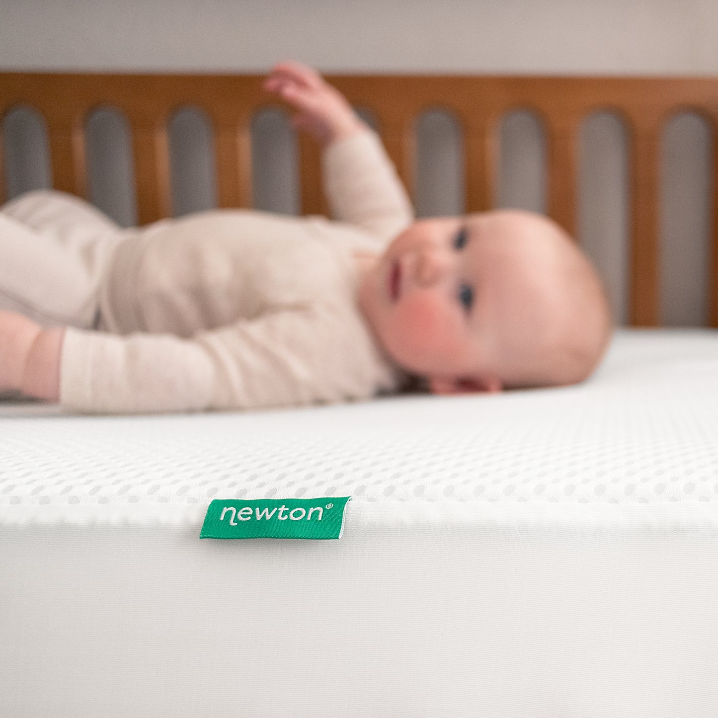 A baby in beige clothing lies on a white crib mattress in a wooden crib, with a green Newton Baby tag visible on the Essential Waterproof Crib Pad.