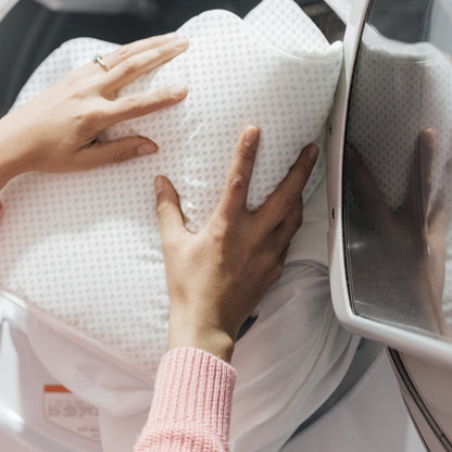A person in a pink sweater puts a white, dotted Essential Waterproof Crib Pad by Newton Baby into the washing machine.