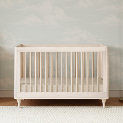 The Babyletto Lolly Convertible Crib, crafted from New Zealand pine wood, features vertical slats and a minimalist design. It stands on a patterned rug, with a striped mattress, against soft blue and white cloud wallpaper.