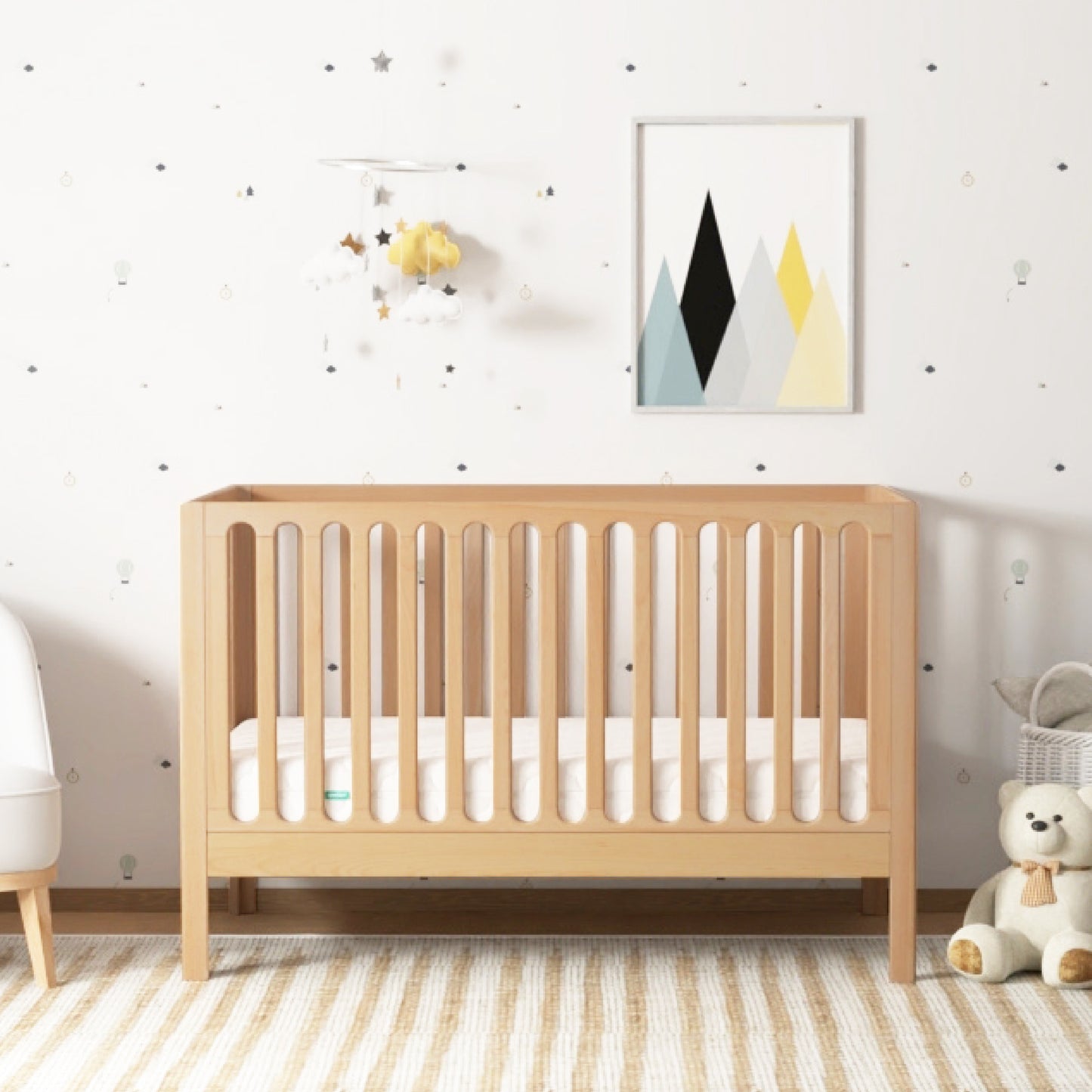 The Newton Galileo Convertible Crib by Newton Baby with white bedding sits in a nursery featuring mountain-themed wall art, a hanging mobile, a teddy bear, and a striped rug. Its design allows easy toddler bed conversion as your child grows. 999-8420-NAT