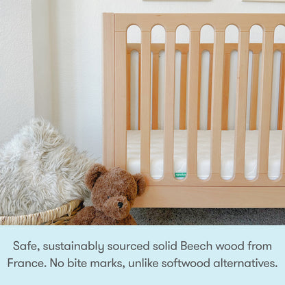 The Newton Galileo Convertible Crib by Newton Baby, made from sustainably sourced French beech wood with safe, bite-proof vertical slats, stands beside a fluffy rug and a brown teddy bear in a basket. Easily converts to a toddler bed.