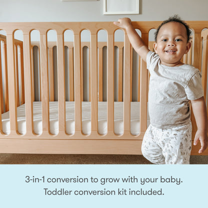 A smiling toddler stands by a Newton Galileo Convertible Crib from Newton Baby in a nursery. Text below reads, 3-in-1 convertible crib grows with your child—toddler bed conversion kit included.