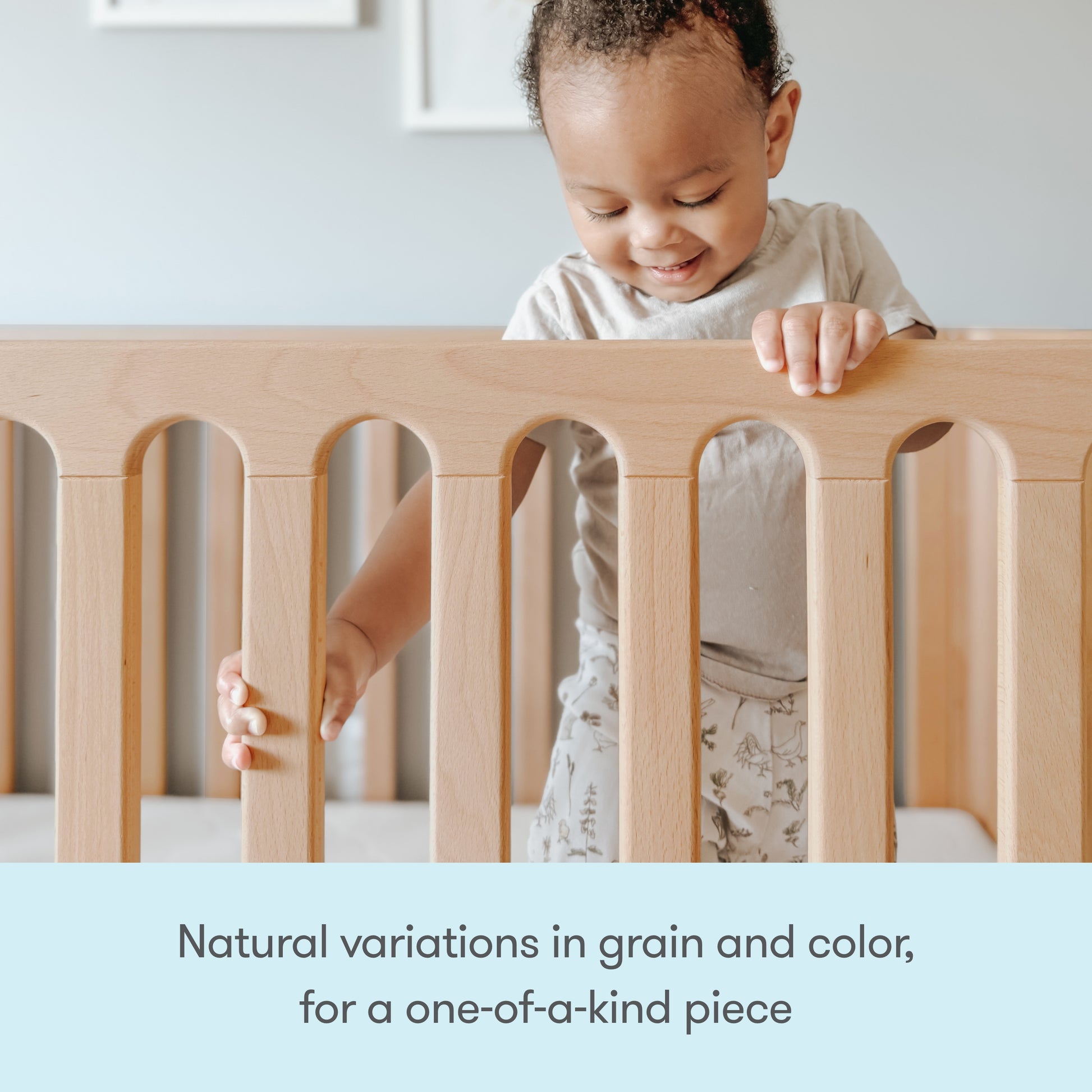 A smiling toddler stands in the Newton Galileo Convertible Crib by Newton Baby, holding the railing. The crib features unique natural grain and color variations, making each one-of-a-kind. Text: Natural variations for a unique convertible crib.