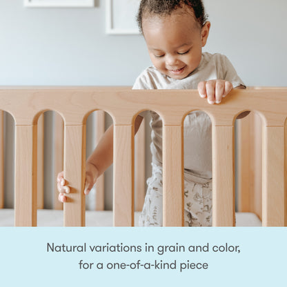 A smiling toddler stands in the Newton Galileo Convertible Crib by Newton Baby, holding the railing. The crib features unique natural grain and color variations, making each one-of-a-kind. Text: Natural variations for a unique convertible crib.