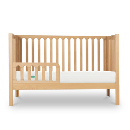 The Newton Galileo Convertible Crib by Newton Baby features a modern beech wood design with vertical slats on three sides and a partial left guard rail. Shown with a white mattress, it easily converts to a toddler bed. Displayed on a white background.