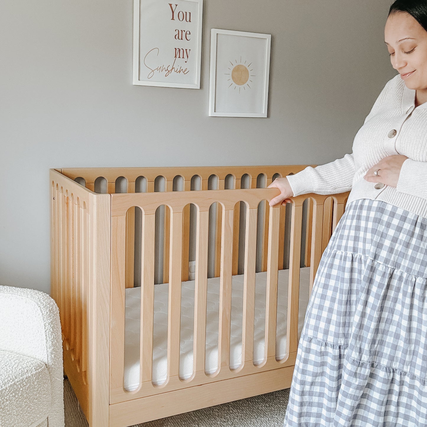 A woman stands beside the Newton Baby Galileo Convertible Crib in a softly lit nursery, gently touching her pregnant belly. Framed prints, including one that says "You are my sunshine," decorate the wall behind her.
