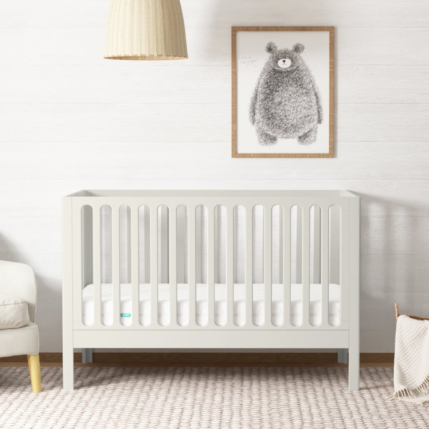 The Newton Galileo Convertible Crib by Newton Baby with a mattress rests on a woven rug in a bright nursery. In the background are a beige armchair, hanging light, and framed bear illustration. 999-8402-WRW