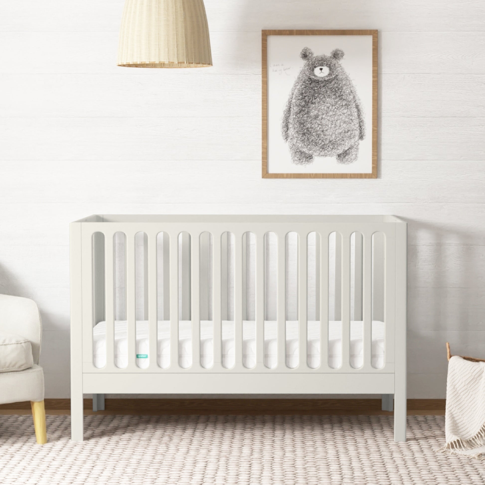 The Newton Galileo Convertible Crib by Newton Baby with a mattress rests on a woven rug in a bright nursery. In the background are a beige armchair, hanging light, and framed bear illustration. 999-8402-WRW