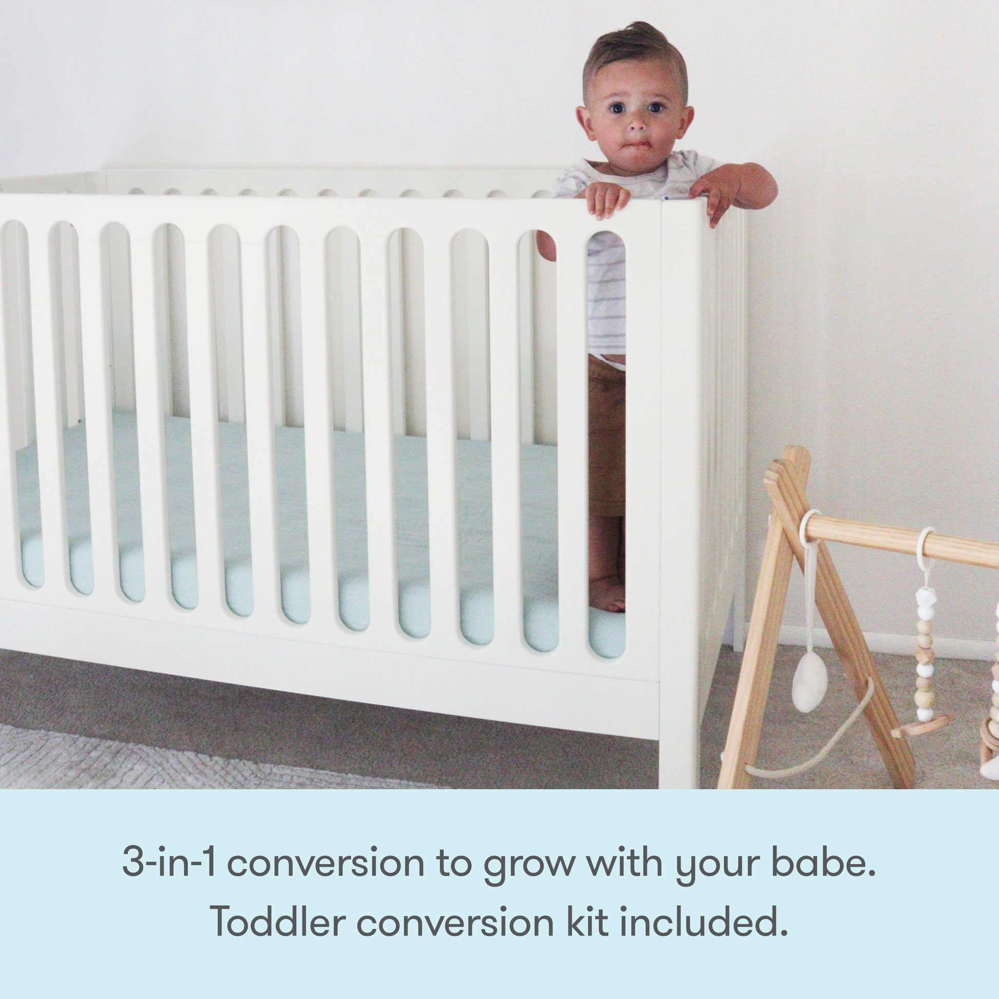 A toddler stands holding the rail of the white Newton Galileo Convertible Crib by Newton Baby. Next to it is a wooden baby gym with hanging toys. Text: 3-in-1 convertible crib grows with your child. Toddler bed conversion kit included.