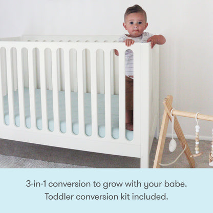 A toddler stands holding the rail of the white Newton Galileo Convertible Crib by Newton Baby. Next to it is a wooden baby gym with hanging toys. Text: 3-in-1 convertible crib grows with your child. Toddler bed conversion kit included.
