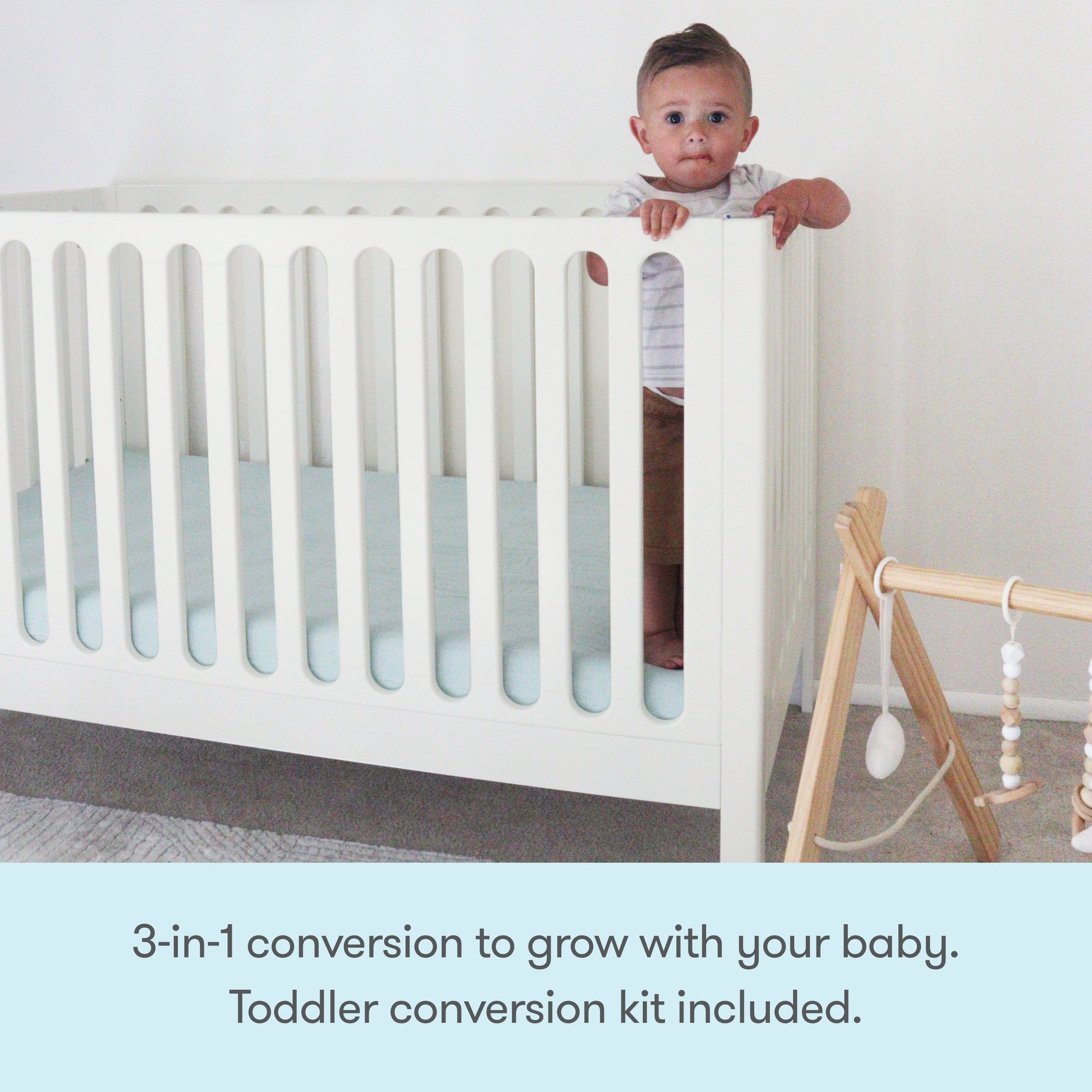 A baby stands holding the side of a Newton Galileo Convertible Crib by Newton Baby, featuring beech wood and a light blue mattress in a minimal nursery. Beside it is a wooden baby gym. Text: 3-in-1 convertible. Toddler kit included.